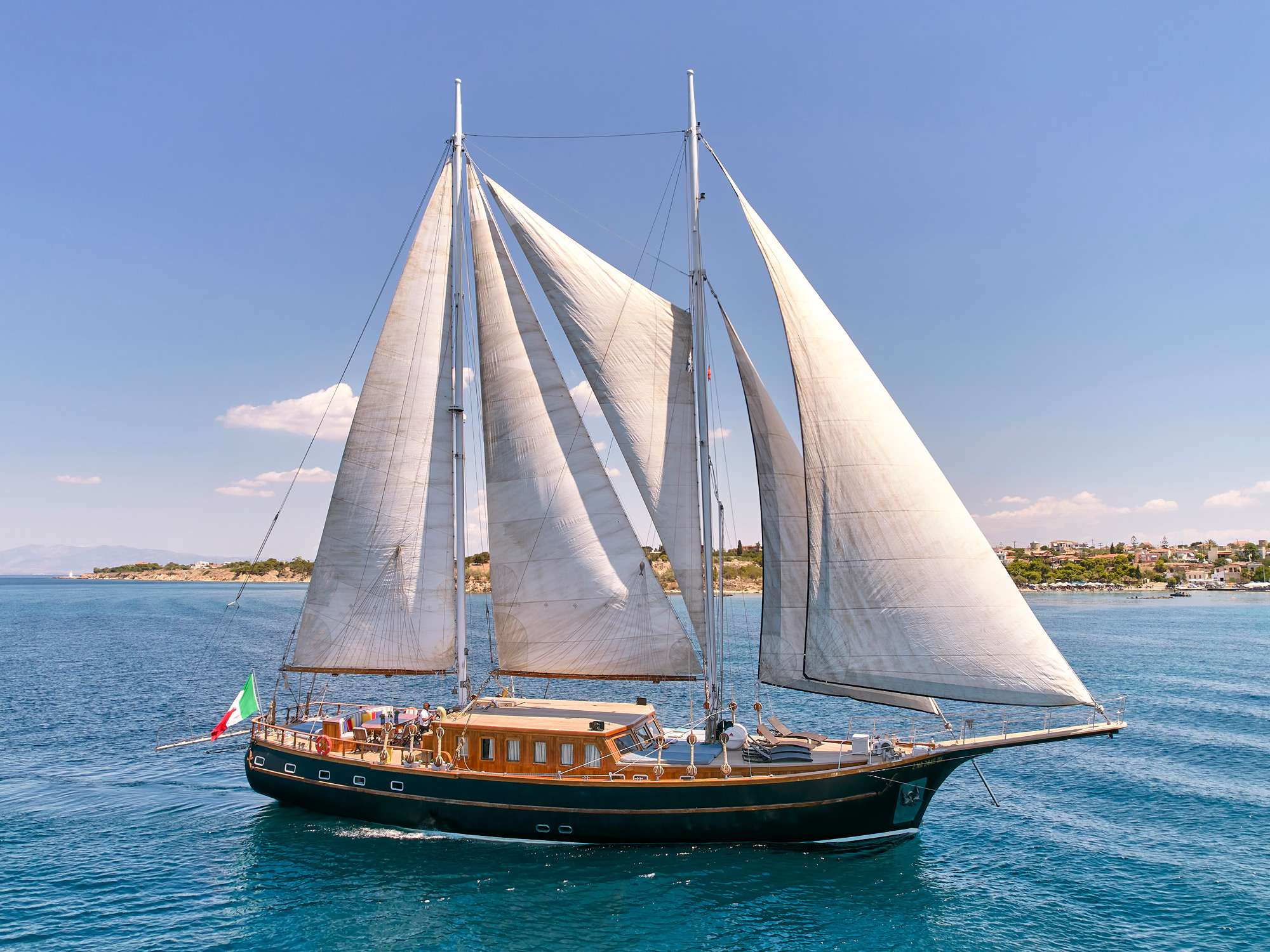 The sailing schooner Myra was designed and built by the Turkish Ege Yat yacht builders in 2003, and has been refitted in 2021/22. Myra is an extremely elegant, classic and refined, the sailing yacht Myra has a traditional vibe about her. Her exterior steel hull is an added value and a unique feature for a schooner. Her beautiful exterior is with gloss varnished bulwarks and teak deck. The interior is spacious and warm boasting fine woods panels and teak-holly inlay floors.
The salon is in the raised deck house, large ports allow ample natural light and great viewing. On the port side 2 formal dining tables seat all 12 guests with room to spare, on the starboard side a built in lounge and coffee table invite guests to relax in comfort. A flat screen TV and audio system is available for indoor entertainment. Four double guest cabins are
forward of the salon, while 2 twin guests cabins are aft of the salon.
Exterior living on charter yacht MYRA is plentiful. The aft deck offers an alfresco dining option, seating and aft facing sunbeds, shade from a Bimini cover can be provided. Additional sunbeds on the foredeck allow everyone to find a place to relax.
The sailing schooner Myra boasts generous wrap around teak decks, with high rails, the yacht is easy to walk around both at anchor and under sail.
Myra is RINA and CE certified.